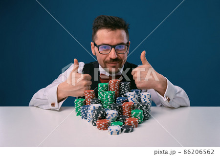 Fellow in glasses, black vest and shirt sitting at white table with stacks of chips on it, posing on blue background. Poker, casino. Close-up. 86206562