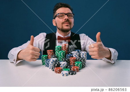 Player in glasses, black vest and shirt sitting at white table with stacks of chips on it, posing on blue background. Poker, casino. Close-up. 86206563