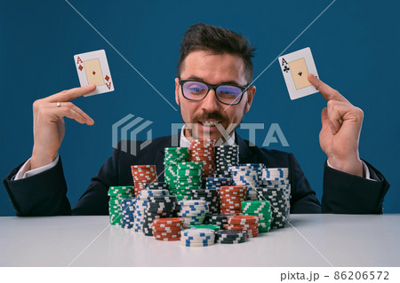 Man in glasses, black suit sitting at white table with stacks of chips, holding two playing cards, posing on blue background. Poker, casino. Close-up. Man in glasses, black suit sitting at white table with stacks of chips, holding two playing cards, posing on blue background. Poker, casino. Close-up. 86206572
