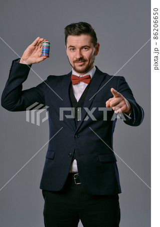 Man in black classic suit and red bow-tie showing some colored chips, posing against gray studio background. Gambling, poker, casino. Close-up. 86206650
