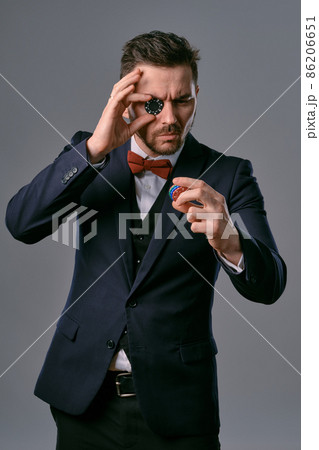 Man in black classic suit and red bow-tie showing some colored chips, posing against gray studio background. Gambling, poker, casino. Close-up. 86206651