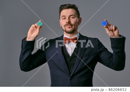 Man in black classic suit and red bow-tie showing some colored chips, posing against gray studio background. Gambling, poker, casino. Close-up. Man in black classic suit and red bow-tie showing some colored chips, posing against gray studio background. Gambling, poker, casino. Close-up. 86206652