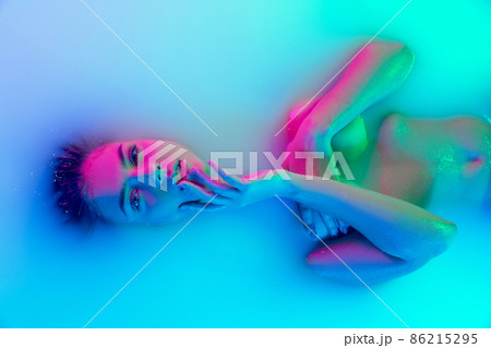 Top view of female face, young girl in the milk bath with soft glowing in blue-pink neon light. Beauty, fashion, style, skincare concept Top view of female face, young girl in the milk bath with soft glowing in blue-pink neon light. Beauty, fashion, style, skincare concept 86215295