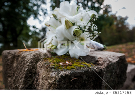 Floral arrangement with white flowers, wedding bouquet with a ribbon and two gold rings, lying on a stone. Wedding decor, artwork, florist 86217568