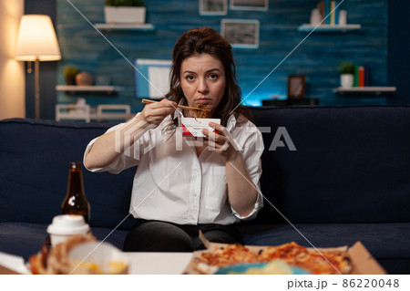 Tired woman holding chopsticks and chinese takeaway noodle box sitting on couch Tired woman holding chopsticks and chinese takeaway noodle box sitting on couch 86220048