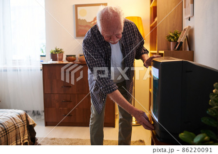 Senior man bending by tv set while switching it on or off Senior man bending by tv set while switching it on or off 86220091