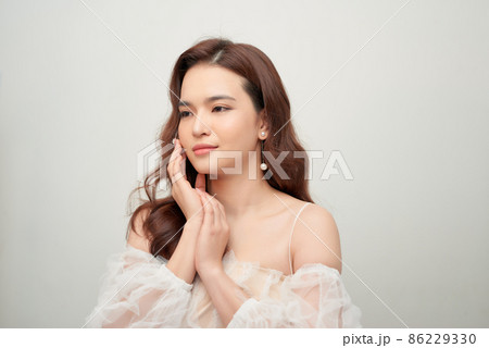 Portrait of a cheerful young woman with long hair dressed in white dress posing isolated over gray background 86229330