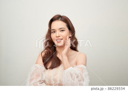 Tender asian woman wearing natural makeup, white blouse, touching her facial skin and gazing at camera Tender asian woman wearing natural makeup, white blouse, touching her facial skin and gazing at camera 86229334