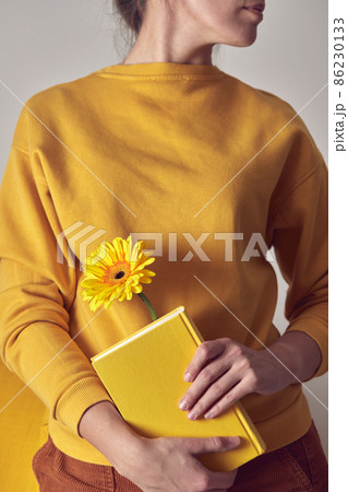 A yellow book with a flower bookmarked in the hands of a woman on a yellow sweater background. Front view. A yellow book with a flower bookmarked in the hands of a woman on a yellow sweater background. Front view. 86230133