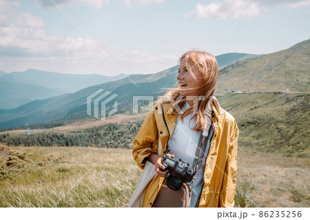 Beautiful smile blonde girl tourist in yellow raincoat and with a camera looks at the beautiful green hills of the Carpathians of Ukraine on a sunny day 86235256