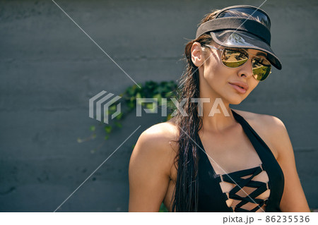 Woman posing sitting on a green grass of a recreation center or hotel with a swimming pool. Dressed in a black swimsuit, sun visor and sunglasses. 86235536