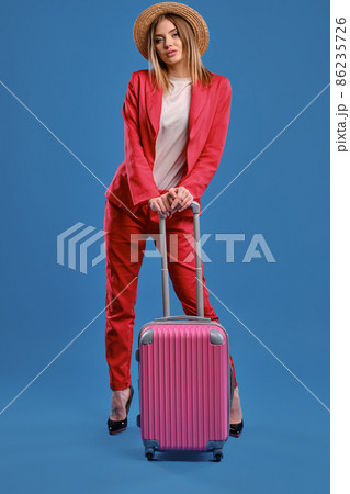 Blonde woman in straw hat, white blouse, red pantsuit, high black heels. She is leaning on a handle of pink suitcase while posing on blue background 86235726