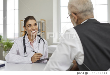 Friendly young female doctor in hospital office consults senior male patient and makes entries in his medical card. Smiling nurse sitting at table talking to mature man sitting with his back to camera 86235744