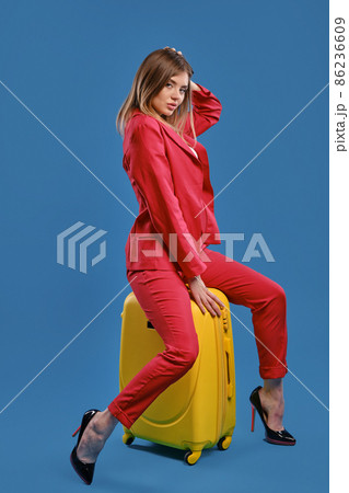 Blonde female in white blouse, red pantsuit, high black heels. She touching hair, sitting on yellow suitcase, posing sideways on blue background 86236609