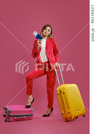 Blonde female in red pantsuit, white blouse, black heels. She put her leg on suitcase, yellow one nearby, holding passport and ticket, pink background Blonde female in red pantsuit, white blouse, black heels. She put her leg on suitcase, yellow one nearby, holding passport and ticket, pink background 86236827