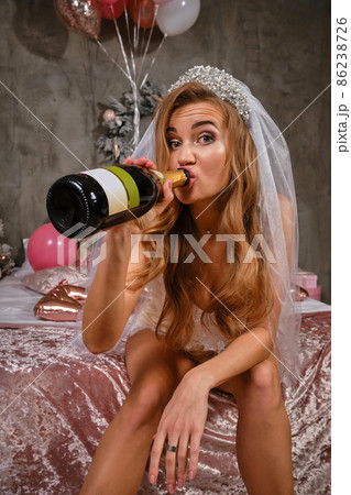 Woman in beige lingerie and white veil enjoying hen-party of bride, sitting on bed, drinking champagne. Christmas decorations, balloons. Close-up. Woman in beige lingerie and white veil enjoying hen-party of bride, sitting on bed, drinking champagne. Christmas decorations, balloons. Close-up. 86238726
