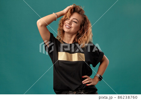 Ginger girl with a curly hair, wearing black t-shirt with a golden stripe is posing standing against a blue studio background. Close-up shot. 86238760
