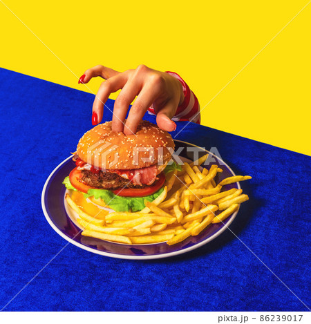 Food pop art photography. Female hand and hamburger, french fries on bright blue tablecloth isolated on yellow background. Vintage, retro style 86239017