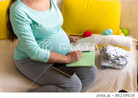 Cute Pregnant Woman writing packing list for maternity hospital With Notebook Prepares bags. Young Ledy In Pregnancy Have Fun Spend Time On Sofa At Home. Motherhood, Medicine Health and Care Concept 86239028