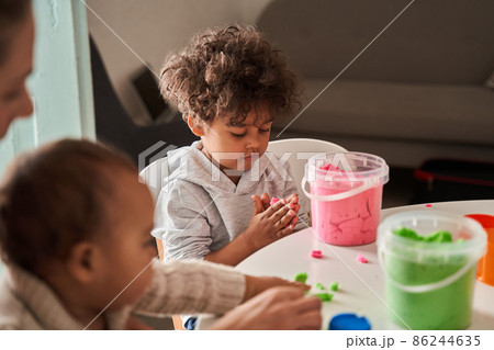 Concentrated brunette boy looking attentively and playing with purple sand on a white table Concentrated brunette boy looking attentively and playing with purple sand on a white table 86244635