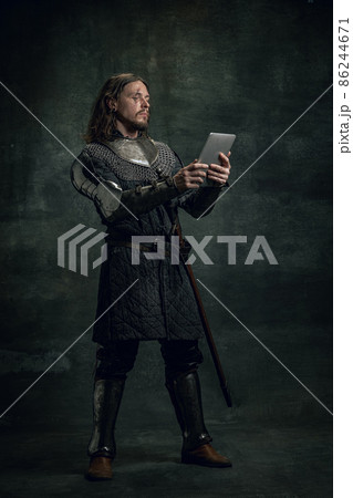 Vintage style portrait of brutal smiling man, medieval warrior or knight with dirty wounded face using modern gadget isolated over dark background. Comparison of eras Vintage style portrait of brutal smiling man, medieval warrior or knight with dirty wounded face using modern gadget isolated over dark background. Comparison of eras 86244671