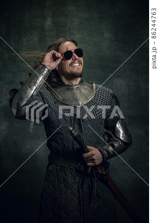 Vintage style portrait of brutal smiling man, medieval warrior or knight with dirty wounded face in sunglasses isolated over dark background. Comparison of eras Vintage style portrait of brutal smiling man, medieval warrior or knight with dirty wounded face in sunglasses isolated over dark background. Comparison of eras 86244763