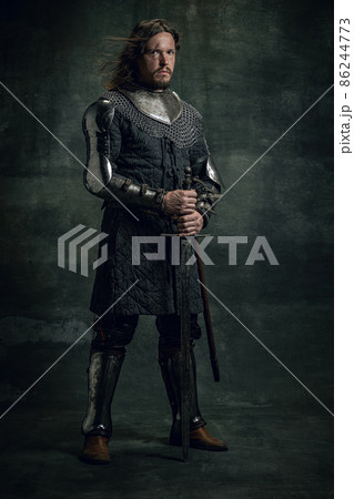 Vintage style portrait of brutal seriuos man, medieval warrior or knight with dirty wounded face holding big sword isolated over dark background. Comparison of eras Vintage style portrait of brutal seriuos man, medieval warrior or knight with dirty wounded face holding big sword isolated over dark background. Comparison of eras 86244773