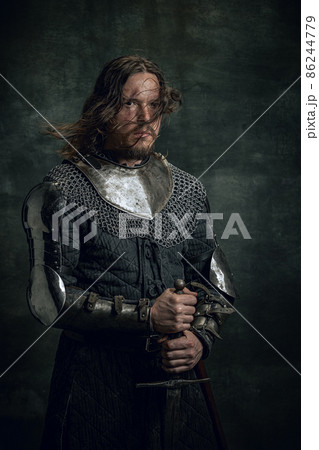 Portrait of medieval warrior or knight with dirty wounded face holding big sword isolated over dark background. Comparison of eras, history 86244779
