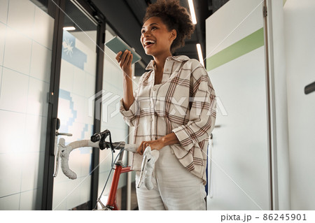 Joyful multiracial woman standing with bike at the hall and recording voice message 86245901
