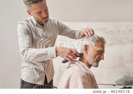 Concentrated mature man shaving with trimmer back of the head of his retirement father 86246578
