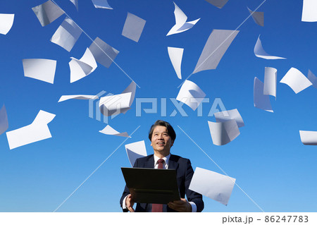 書類が舞う青空のもとでパソコンを手に持つスーツ姿の男性 86247783