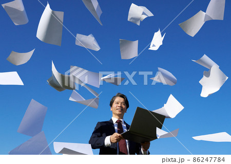 書類が舞う青空のもとでパソコンを手に持つスーツ姿の男性 86247784