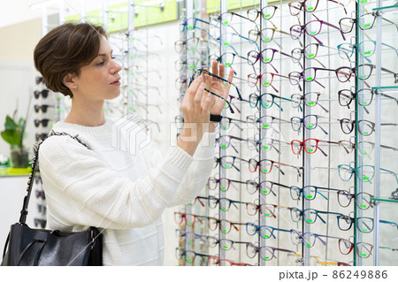 Photo of young thinking attractive brunette woman in a white sweater chooses glasses for vision on a Photo of young thinking attractive brunette woman in a white sweater chooses glasses for vision on a 86249886