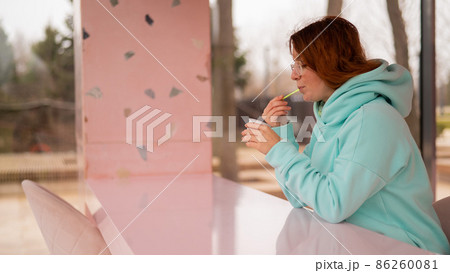 Caucasian red-haired woman eats ice cream with a plastic spoon while sitting alone in a cafe. Caucasian red-haired woman eats ice cream with a plastic spoon while sitting alone in a cafe. 86260081