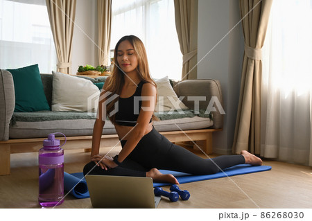 Smiling asian woman doing yoga on fitness mat in living room and watching video tutorial on laptop compute. 86268030