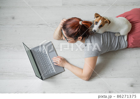 A European red-haired woman is lying on the floor with her dog and is typing on a laptop. Female student studying remotely at home in quarantine. A loyal puppy Jack Russell Terrier. Exam preparation. 86271375