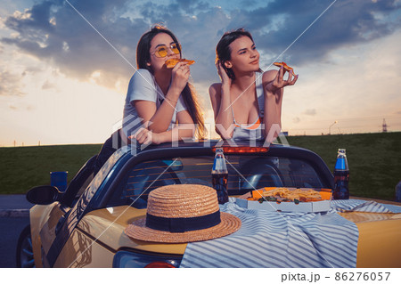 Charming ladies in casual outfit are eating pizza, posing in yellow car with french fries, hat and soda in glass bottles on trunk. Fast food. Mock up 86276057