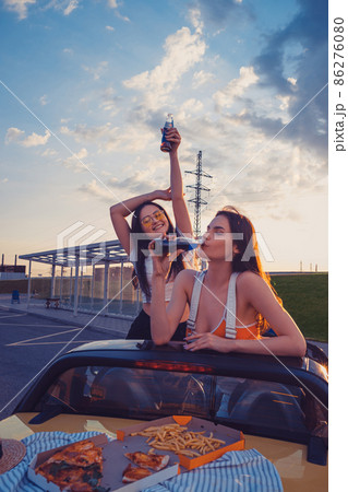 Happy young girls are having fun and drinking soda, posing in yellow car cabrio with french fries and pizza on its trunk. Fast food. Copy space Happy young girls are having fun and drinking soda, posing in yellow car cabrio with french fries and pizza on its trunk. Fast food. Copy space 86276080