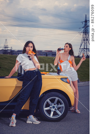 Attractive models in casual outfit eating pizza, enjoying soda water in glass bottles, posing near yellow car on parking lot. Full length, copy space Attractive models in casual outfit eating pizza, enjoying soda water in glass bottles, posing near yellow car on parking lot. Full length, copy space 86276099