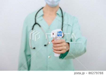 Temperature measurement gun in doctor hands. Close-up shot of doctor wearing protective surgical mask ready to use infrared isometric thermometer gun to check body temperature for virus symptoms. Temperature measurement gun in doctor hands. Close-up shot of doctor wearing protective surgical mask ready to use infrared isometric thermometer gun to check body temperature for virus symptoms. 86279766