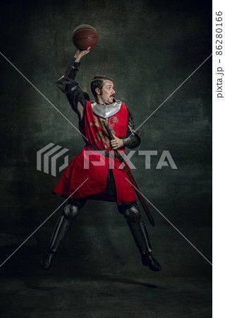 Portrait of medieval warrior or knight with dirty wounded face playing with basketball ball isolated over dark background. Comparison of eras, history Portrait of medieval warrior or knight with dirty wounded face playing with basketball ball isolated over dark background. Comparison of eras, history 86280166