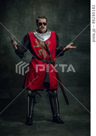 Surprised young man in image of medieval warrior or knight with dirty wounded face using phone isolated over dark background. Comparison of eras Surprised young man in image of medieval warrior or knight with dirty wounded face using phone isolated over dark background. Comparison of eras 86280182