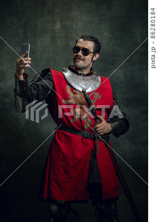 Smiling young man in image of medieval warrior or knight with dirty wounded face using phone isolated over dark background. Comparison of eras Smiling young man in image of medieval warrior or knight with dirty wounded face using phone isolated over dark background. Comparison of eras 86280184