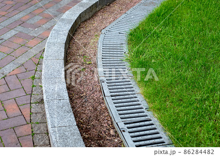 iron drainage grate on the roadside with green grass and stone pebbles at the granite curb along the pedestrian pavement of brick tiles, nobody. iron drainage grate on the roadside with green grass and stone pebbles at the granite curb along the pedestrian pavement of brick tiles, nobody. 86284482