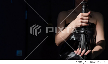 BDSM concept. A woman in a leather dress holds a black whip with a long fringe. Role-playing games for adults. Close-up of sex toys for domination. 86294203