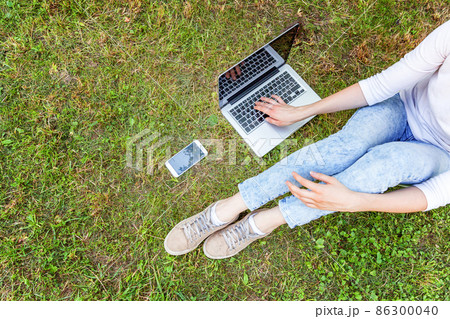 Freelance business concept. Woman legs on green grass lawn in city park, hands working on laptop pc computer. Lifestyle authentic candid student girl studying outdoors. Mobile Office 86300040