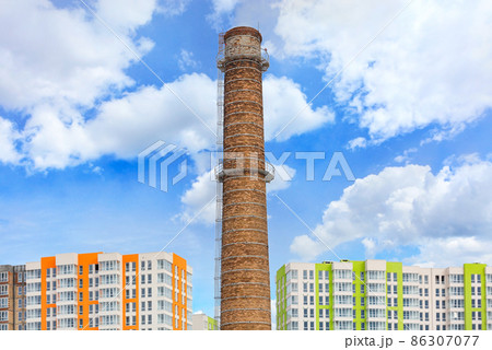 The brick chimney of the thermal power plant rises above the new houses under construction in the sleeping area of the city against the backdrop of a blue sky and white clouds. 86307077