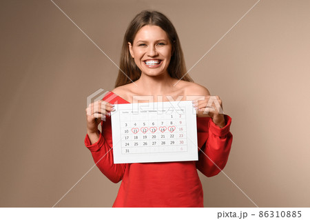 Excited young woman holding calendar with marked menstrual cycle days on beige background 86310885