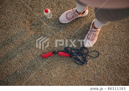 High angle view of female athlete legs wearing pink sneakers and standing on an asphalt treadmill next to a lying down skipping rope and water bottle 86311163