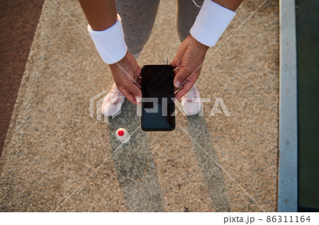 Top view of female athlete hands in white terry wristbands holding a smartphone with empty blank black screen and browsing on websites and mobile applications. Top view of female athlete hands in white terry wristbands holding a smartphone with empty blank black screen and browsing on websites and mobile applications. 86311164